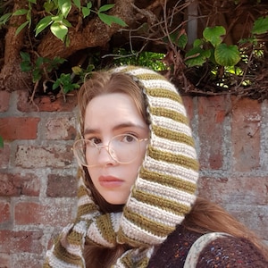 May include: A crochet hood scarf featuring alternating vertical stripes in olive green and cream. The accessory is constructed with a ribbed texture and includes long ends that drape down the sides, framing the face with a soft, knit appearance.