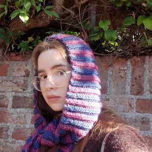 May include: A crochet hood scarf featuring horizontal stripes in alternating shades of deep magenta and dark blue. The accessory is constructed with a ribbed texture and drapes around the head and neck, providing coverage with its long, integrated ends.