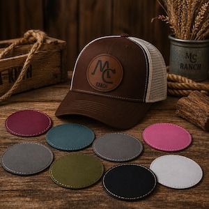 Könnte beinhalten: Eine braune Baseballkappe mit Mesh-Rückseite und einem Lederaufnäher mit der Aufschrift "MC RANCH". Das Bild zeigt auch mehrere runde Untersetzer in verschiedenen Farben, darunter Burgunderrot, Türkis, Grau, Olivgrün, Schwarz, Rosa und Hellgrau.