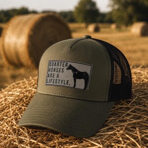 Könnte beinhalten: Eine olivgrüne Trucker-Cap mit schwarzem Netzrücken und einem rechteckigen Aufnäher auf der Vorderseite. Der Aufnäher zeigt die schwarze Silhouette eines Pferdes und den Text QUARTER HORSES ARE A LIFESTYLE in schwarzer Schrift auf grauem Grund.