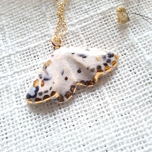 May include: A gold-colored chain necklace with a pendant shaped like a butterfly wing. The pendant is white with black and gold accents. The necklace is displayed on a white textured surface.