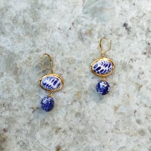 May include: A pair of gold-toned earrings. Each earring features an oval-shaped top with a blue and white pattern, connected to a dangling blue sphere. The earrings are displayed against a light-colored, textured background.