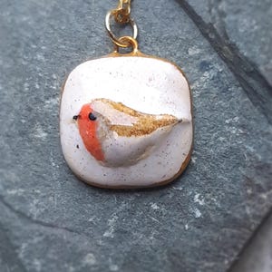 K&ouml;nnte beinhalten: Eine goldene Kettenkette mit einem wei&szlig;en Keramik-Anh&auml;nger mit einem handbemalten Rotkehlchen. Das Rotkehlchen hat eine rote Brust, braune Fl&uuml;gel und ein schwarzes Auge.