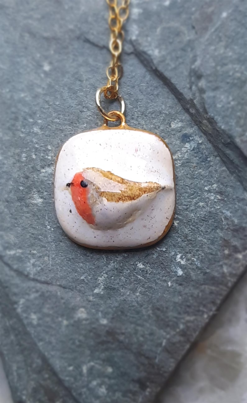 K&ouml;nnte beinhalten: Eine goldene Kettenkette mit einem wei&szlig;en Keramik-Anh&auml;nger mit einem handbemalten Rotkehlchen. Das Rotkehlchen hat eine rote Brust, braune Fl&uuml;gel und ein schwarzes Auge.