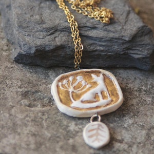 May include: A gold chain necklace with a white ceramic pendant featuring a stylized deer and a tree. The pendant is attached to a smaller white ceramic charm with a tree design.