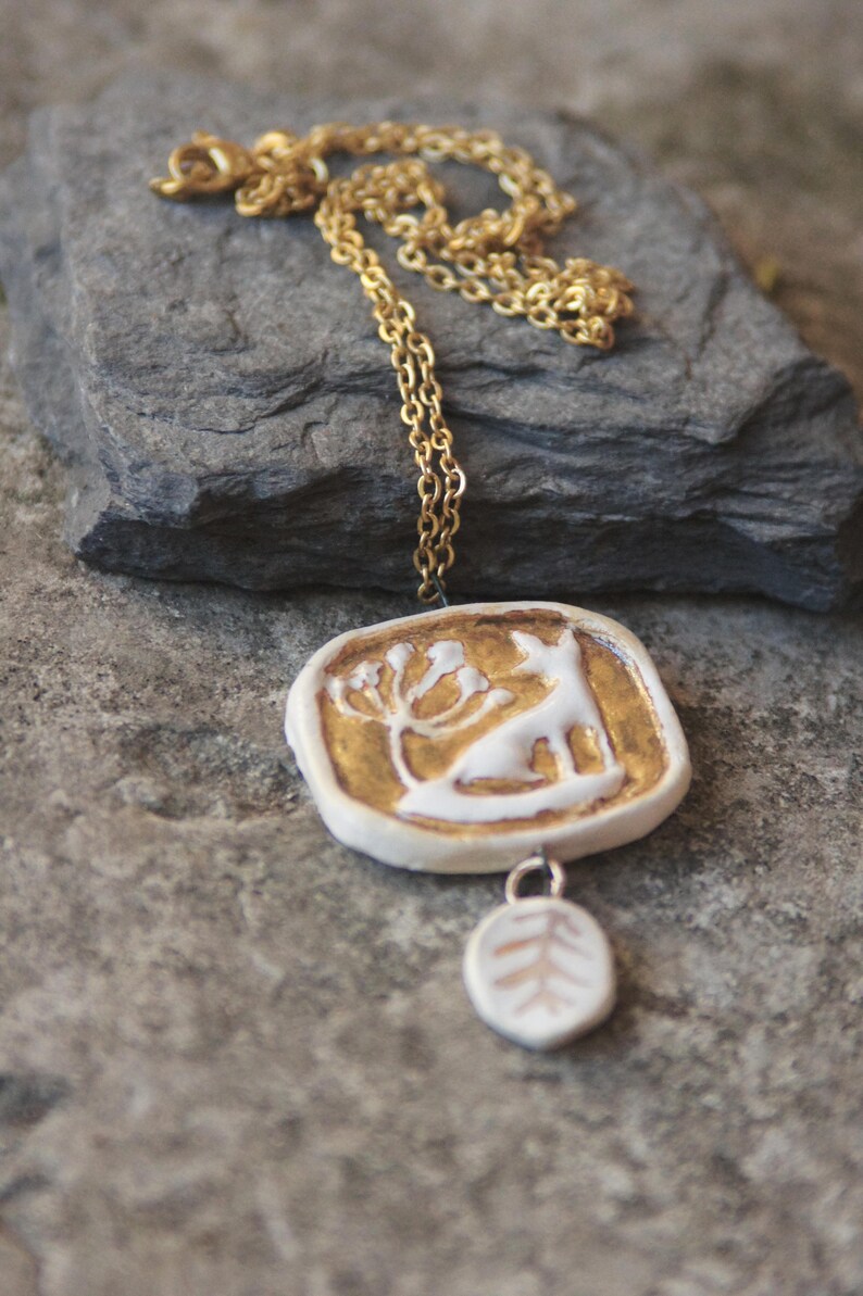 May include: A gold chain necklace with a white ceramic pendant featuring a stylized deer and a tree. The pendant is attached to a smaller white ceramic charm with a tree design.