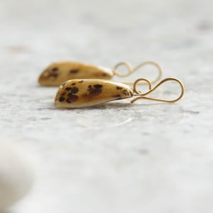 Peut inclure: Une paire de boucles d'oreilles pendantes avec des breloques lisses en forme de goutte, dotées d'une base couleur crème et de motifs tachetés brun foncé irréguliers. Les breloques sont fixées à des crochets en métal doré courbés.