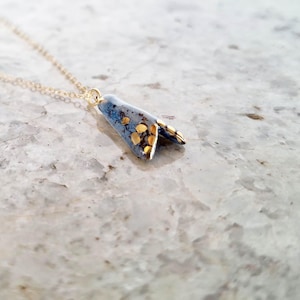 Könnte beinhalten: Eine zarte Halskette mit einem kleinen, gefalteten Anhänger in blau-grauer Sprenkeloptik, der mit verstreuten goldfarbenen Flecken akzentuiert ist. Der Anhänger hängt an einer dünnen, hellen Metallkette, die an der oberen Spitze befestigt ist.