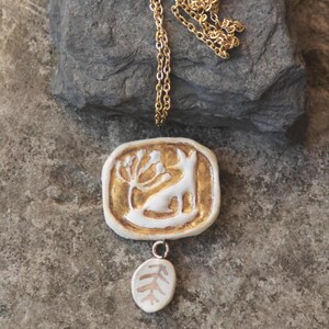 May include: A gold chain necklace with a white ceramic pendant featuring a stylized animal and a leaf design. The pendant is accented with gold paint.