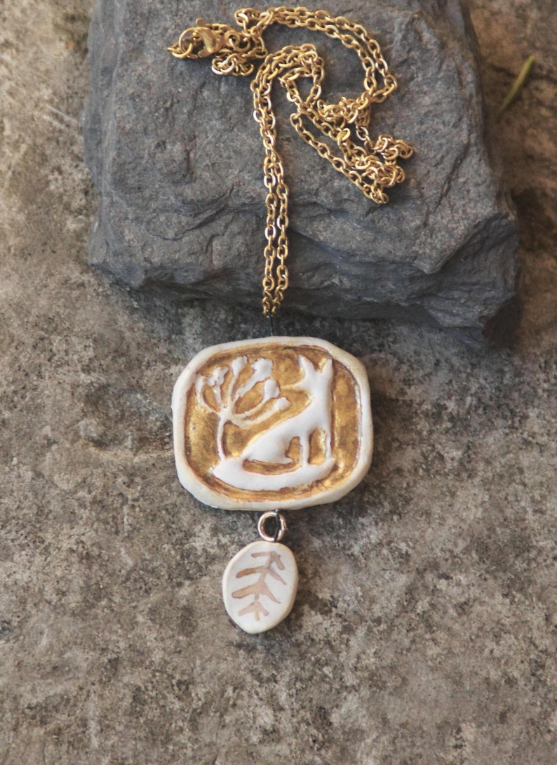 May include: A gold chain necklace with a white ceramic pendant featuring a stylized animal and a leaf design. The pendant is accented with gold paint.