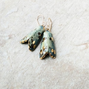 May include: A pair of handcrafted moth-shaped earrings. The earrings are teal with dark brown spots and gold leaf accents. They have gold-coloured ear wires. The earrings are displayed on a light grey surface.