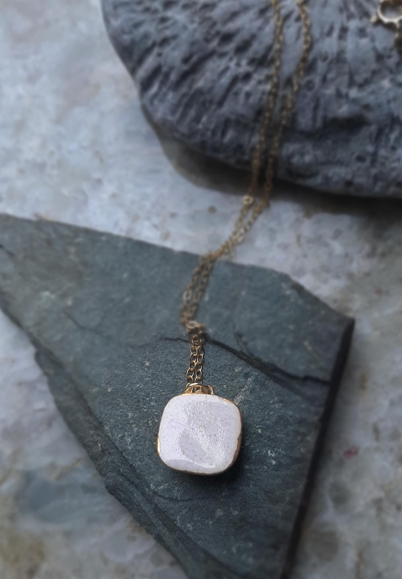 K&ouml;nnte beinhalten: Ein quadratischer Anh&auml;nger aus wei&szlig;er Keramik mit einer goldenen Kette. Der Anh&auml;nger hat eine strukturierte Oberfl&auml;che und h&auml;ngt an einer goldenen Kette.