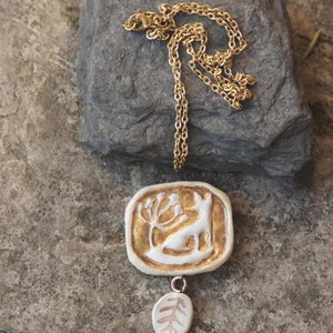 May include: A gold chain necklace with a white ceramic pendant featuring a stylized deer with a tree. The pendant has a smaller white ceramic charm hanging from it, shaped like a leaf.
