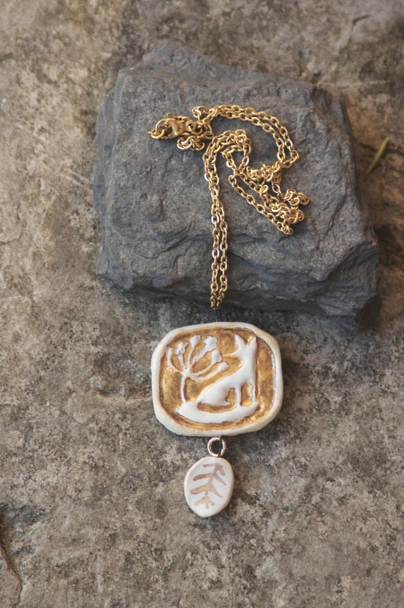 May include: A gold chain necklace with a white ceramic pendant featuring a stylized deer with a tree. The pendant has a smaller white ceramic charm hanging from it, shaped like a leaf.