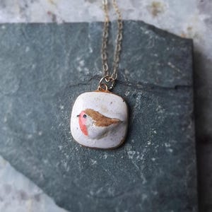 K&ouml;nnte beinhalten: Eine Halskette mit einem wei&szlig;en Keramik-Anh&auml;nger und einer goldenen Kette. Der Anh&auml;nger zeigt einen handbemalten Rotkehlchen mit braunen und roten Akzenten.