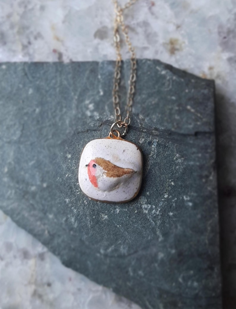 K&ouml;nnte beinhalten: Eine Halskette mit einem wei&szlig;en Keramik-Anh&auml;nger und einer goldenen Kette. Der Anh&auml;nger zeigt einen handbemalten Rotkehlchen mit braunen und roten Akzenten.