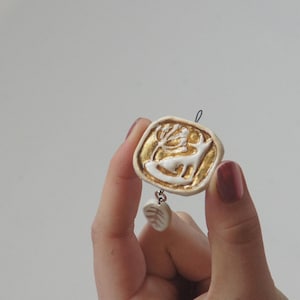 May include: A white ceramic pendant with a gold deer design and a small white ceramic charm hanging from it.
