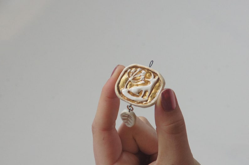 May include: A white ceramic pendant with a gold deer design and a small white ceramic charm hanging from it.