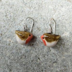 Può includere: Un paio di orecchini artigianali a forma di uccello. Ogni orecchino presenta un corpo marrone e bianco con un petto rosso e un piccolo occhio nero. Gli orecchini hanno ganci per le orecchie color argento.
