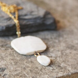 May include: A white ceramic necklace with a gold chain. The necklace features a large, flat, square-shaped pendant with a smaller, oval-shaped pendant hanging from it.