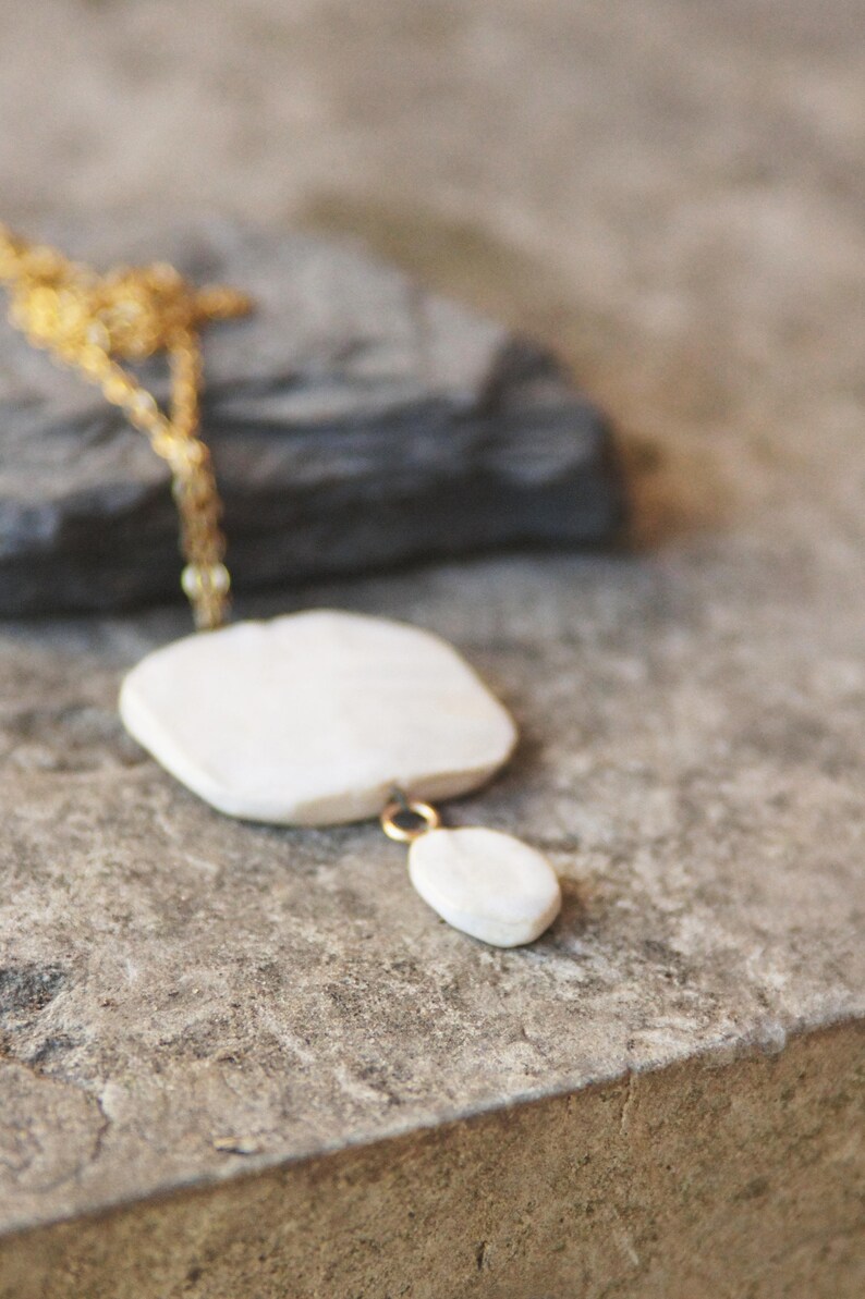 May include: A white ceramic necklace with a gold chain. The necklace features a large, flat, square-shaped pendant with a smaller, oval-shaped pendant hanging from it.