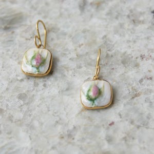 Può includere: Un paio di orecchini pendenti quadrati con un bocciolo di rosa dipinto a mano su base in ceramica bianca, incorniciati in metallo dorato con ganci abbinati per una chiusura sicura e una finitura lucida.