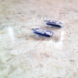 Pode incluir: Um par de brincos retangulares pendentes com uma base de vidro azul profundo e um delicado padrão floral branco. Cada brinco está preso a um gancho de metal prateado simples, proporcionando um visual limpo e polido a estes acessórios.