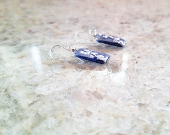 May include: A pair of rectangular drop earrings featuring a deep blue glass base with a delicate white floral pattern. Each earring is attached to a simple silver-toned metal hook wire, providing a clean and polished look for these small accessories.