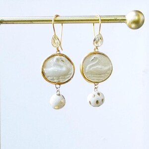 Könnte beinhalten: Ein Paar goldfarbene Ohrhänger. Jeder Ohrring hat einen kreisförmigen Anhänger mit einem Schwanenmotiv, einen tropfenförmigen Akzent und eine baumelnde weiße Perle mit Goldflecken. Die Ohrringe werden auf einer goldenen Stange präsentiert.