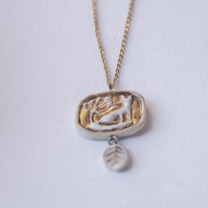 May include: A gold chain necklace with a white ceramic pendant featuring a deer and a leaf design. The pendant is accented with gold paint.