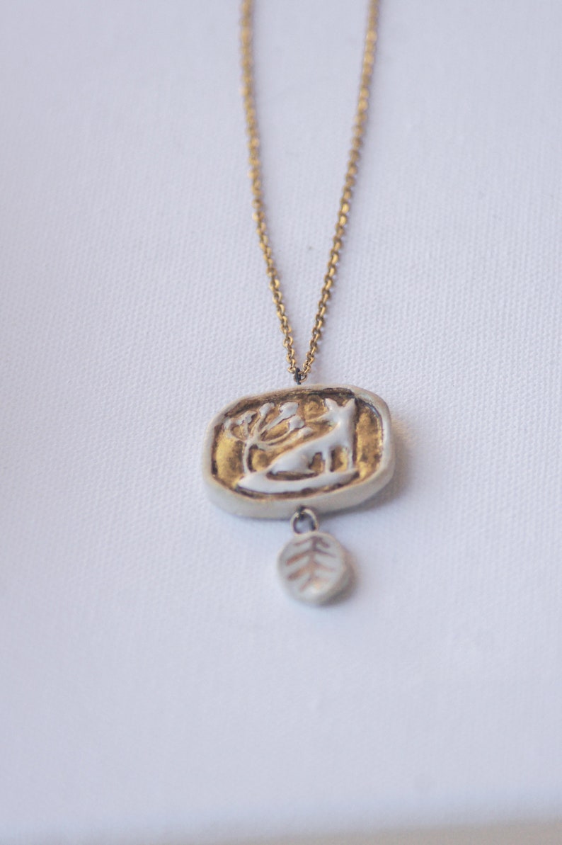 May include: A gold chain necklace with a white ceramic pendant featuring a deer and a leaf design. The pendant is accented with gold paint.
