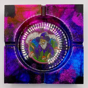 May include: A square resin ashtray with a galaxy-like design in shades of pink, blue, and black. The center features a round, clear insert with a cartoon image of a wizard smoking a pipe and surrounded by mushrooms.