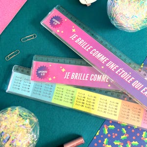 May include: Two pink and clear plastic rulers with the text "Je brille comme une étoile qui explose" and "Je brille comme" printed on them. The rulers have multiplication tables printed on them in different colors.