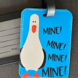 May include: A blue luggage tag with a cartoon seagull illustration. The seagull is white with black accents, a red beak and feet. The tag has the word "MINE!" repeated multiple times in black text.