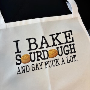 May include: White apron with black text that reads "I BAKE SOURDOUGH AND SAY FUCK A LOT." Two cartoon sourdough bread illustrations are between the words "SOURDOUGH". The apron has a white neck strap and black adjustable buckle.