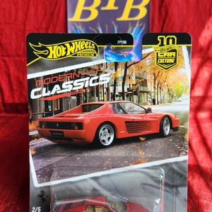 May include: A Hot Wheels die-cast model of a red Ferrari Testarossa, part of the Modern Classics series. The packaging features the Hot Wheels logo and the text "10 Car Culture". The model car is approximately 3 inches long.
