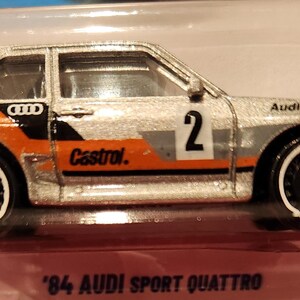 May include: Silver and orange '84 Audi Sport Quattro die-cast model car. The car features the number 2, the word "Castrol", and "Quattro" printed on the side. The car is in a clear plastic package.