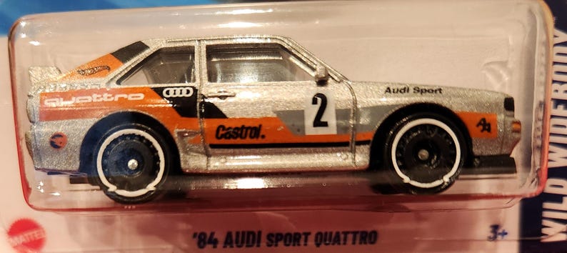 May include: Silver and orange '84 Audi Sport Quattro die-cast model car. The car features the number 2, the word "Castrol", and "Quattro" printed on the side. The car is in a clear plastic package.