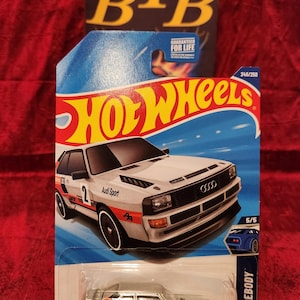 May include: A Hot Wheels die-cast model of an Audi Sport Quattro in a white, orange, and black color scheme. The car has the number 2 on the side and is labeled "Wild Widebody". The packaging includes the Hot Wheels logo and "Guaranteed for Life" text.