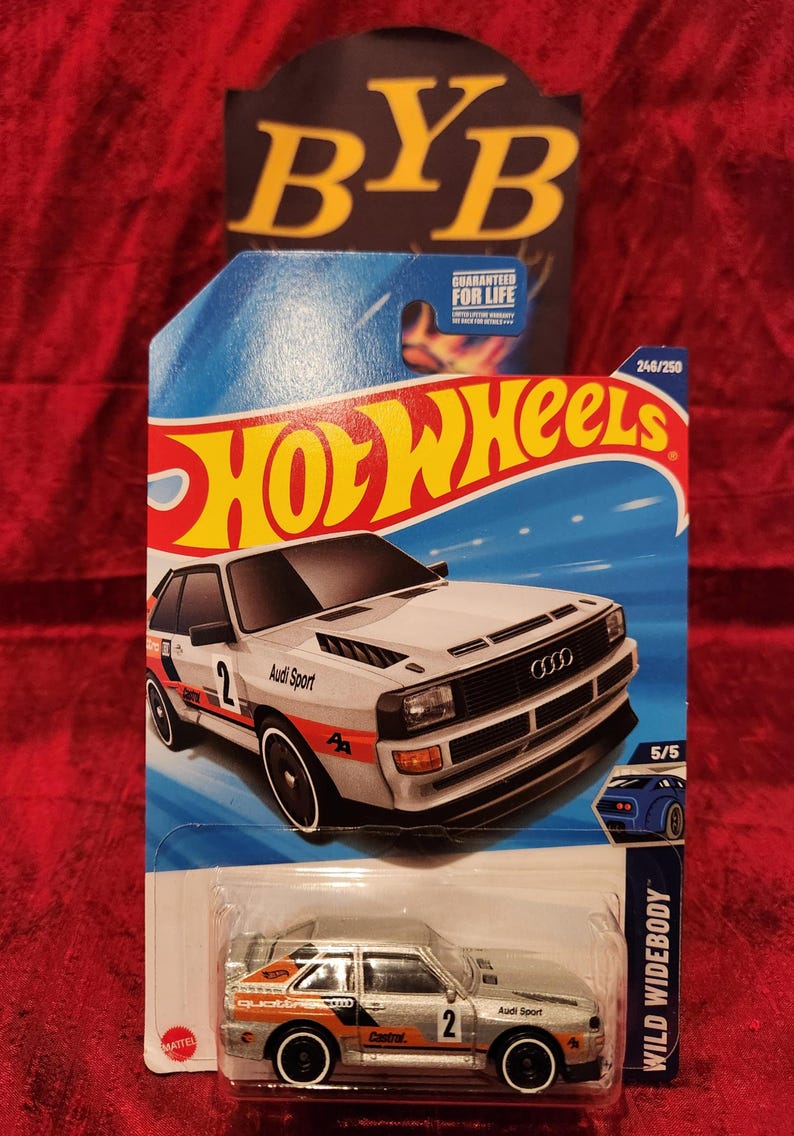 May include: A Hot Wheels die-cast model of an Audi Sport Quattro in a white, orange, and black color scheme. The car has the number 2 on the side and is labeled "Wild Widebody". The packaging includes the Hot Wheels logo and "Guaranteed for Life" text.