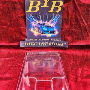 May include: A clear plastic diecast car display case sits in front of a sign with the text "BYB" in yellow letters. The sign also features a blue sports car graphic with lightning bolts and the text "VINTAGE - CUSTOM - DIECAST" and "DIECAST 2DIE4".