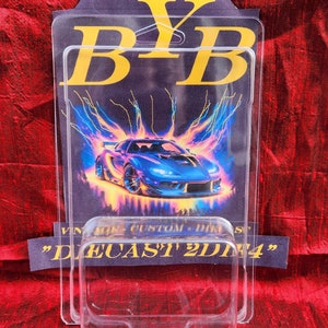 May include: A clear plastic display case with a blue sports car graphic. The graphic has a lightning bolt design and the text "BYB" in gold. The bottom of the case has the text "DIECAST 2DNA".