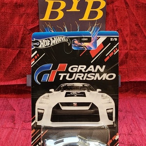 May include: A white Hot Wheels die-cast toy car of a Nissan GT-R Nismo GT3, with the Gran Turismo Academy logo on the front. The car is on a red, white, and black card with the words "Gran Turismo" and "Hot Wheels" printed on it.
