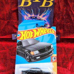 May include: A black 1989 Mercedes-Benz 560 SEC AMG Hot Wheels die-cast car in a plastic blister pack. The car has a black body, black wheels, and a silver grill. The packaging has a blue background with the Hot Wheels logo and the car's name and year.