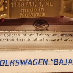 May include: A close-up of a toy car with the text "VOLKSWAGEN "BAJA BU" printed on the bottom. The car is green and has a red stripe on the top. The text "1186 MJ, 1, NL made in Malaysia" is printed on the side of the car.