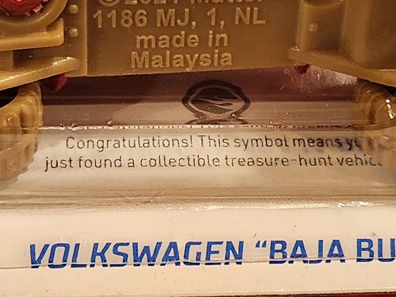 May include: A close-up of a toy car with the text "VOLKSWAGEN "BAJA BU" printed on the bottom. The car is green and has a red stripe on the top. The text "1186 MJ, 1, NL made in Malaysia" is printed on the side of the car.