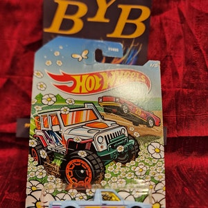 May include: A blue and white Volkswagen Caddy toy car on a white card with a white and orange Jeep toy car in the background. The card has a white daisy pattern and the text "VOLKSWAGEN CADDY" is printed on the bottom.