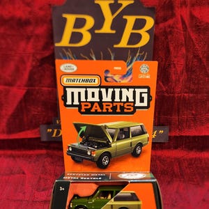 May include: A green Matchbox Moving Parts 1975 Range Rover toy car with an opening hood. The car is made of recycled metal and is part of a toy car collection.