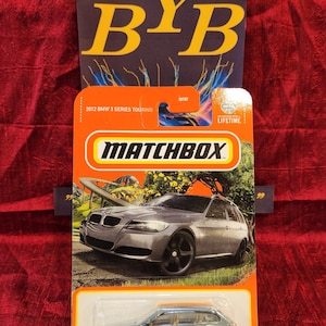 Puede incluir: Un coche de juguete Matchbox, modelo 2012 BMW 3 Series Touring, de color gris, con ruedas negras. El embalaje es naranja y blanco, con el logotipo de Matchbox y el texto "2012 BMW 3 Series Touring".