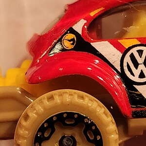 May include: Red, black and white toy car with a Volkswagen logo. The car has a yellow and black stripe on the side and a yellow and black decal on the hood. The car has a black and tan tyre.